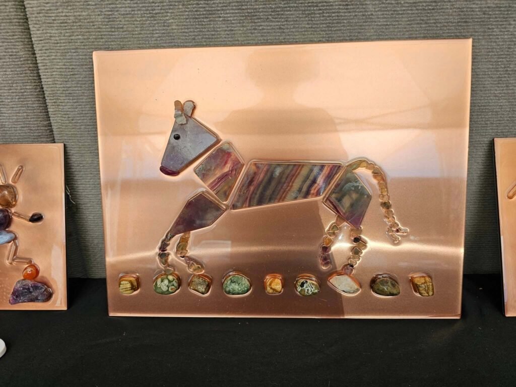 Handcrafted donkey artwork by Therese Tice Art featuring colorful polished stones arranged on a copper-toned background, displayed at an art showcase.