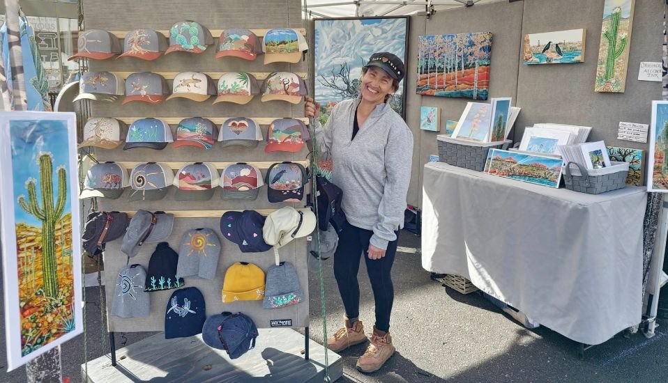 Therese Tice standing beside a display of her hand-painted hats and artwork at an outdoor art show.